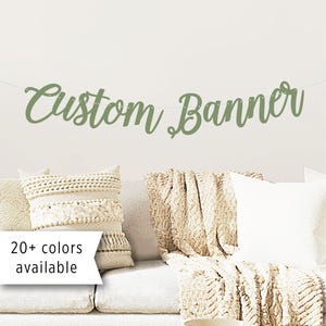 Op de afbeelding: Een saliegroene "Custom Banner" hangt boven een witte bank met decoratieve kussens en een deken. De banner is geregen aan een dun wit touwtje. Een witte banner met de tekst "20+ kleuren beschikbaar" staat in de linker benedenhoek.