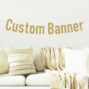 May include: Gold glitter banner with the text "Custom Banner" hanging on a white wall above a white couch with throw pillows.