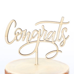Puede incluir: Topper de pastel de madera con la palabra "Congrats" escrita en letra cursiva.