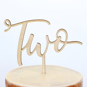 May include: Wooden cake topper with the word "Two" written in a cursive script. The topper is on a stick and is ready to be placed on a cake.