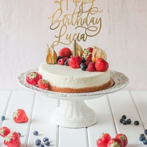 May include: A white cheesecake topped with fresh berries and a gold cake topper that reads "Happy Birthday Lucia".