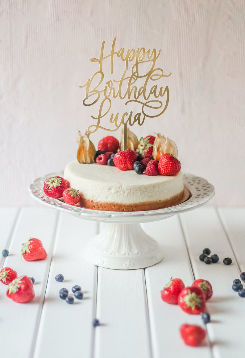 May include: A white cheesecake topped with fresh berries and a gold cake topper that reads "Happy Birthday Lucia".