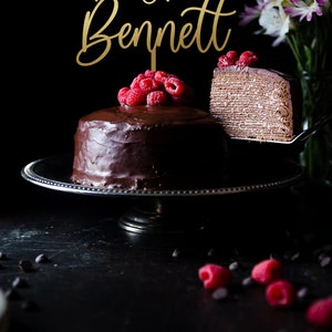 May include: A two-tier chocolate cake with a gold topper that says "Mr. & Mrs. Bennett" and raspberries on top. A slice of the cake is being lifted off the cake stand.
