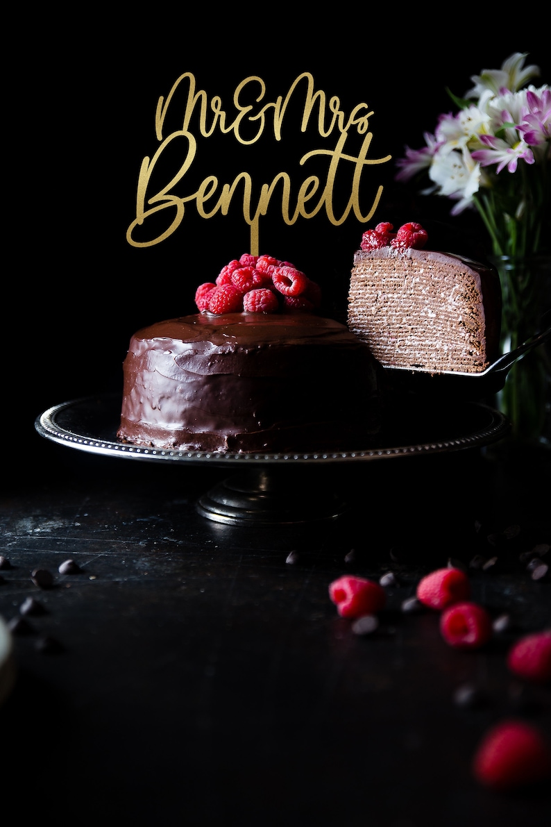 May include: A two-tier chocolate cake with a gold topper that says "Mr. & Mrs. Bennett" and raspberries on top. A slice of the cake is being lifted off the cake stand.