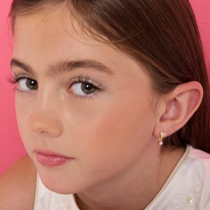 Puede incluir: Un pendiente de tono dorado que presenta un pequeño perno en forma de corazón incrustado con diminutas piedras transparentes, del cual cuelga una sola piedra preciosa redonda de color rosa. El pendiente se lleva en el lóbulo de la oreja.