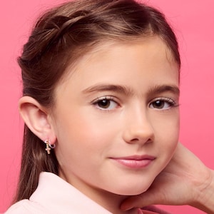 May include: A young person with brown hair and eyes, wearing a light pink shirt, smiles at the camera. They are wearing small gold hoop earrings with a cross charm. The background is a solid pink.