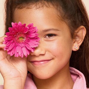 May include: A person smiles, holding a bright pink gerbera daisy in front of one eye. The person wears a pink shirt and has a small earring with a roller skate design. The background is a soft, neutral tone.
