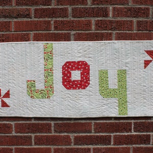 Peut inclure: Une tenture murale matelassée avec le mot "JOY" en lettres de tissu vertes et rouges. Le "J" et le "Y" sont verts avec un motif de lettres, le "O" est rouge avec des pois blancs. Des accents de moulinets rouges et blancs sont à gauche et à droite.