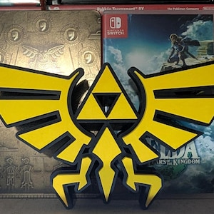 May include: A yellow and black 3D-printed Hylian Crest from The Legend of Zelda, in front of a gold-coloured game case and a Nintendo Switch game case. The game case is for "The Legend of Zelda: Tears of the Kingdom".