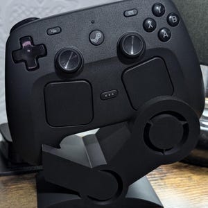 May include: A black Steam Deck controller on a black stand. The controller features various buttons, including a D-pad, analogue sticks, and face buttons. The stand is designed to hold the controller at an angle for display or storage.