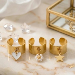 Könnte beinhalten: Drei goldfarbene Ringe mit Anhängern. Einer hat ein weißes Herz, ein anderer eine Ansammlung von Perlen und der dritte einen Stern. Die Ringe liegen auf einer Marmoroberfläche mit einem weißen Tuch im Hintergrund.