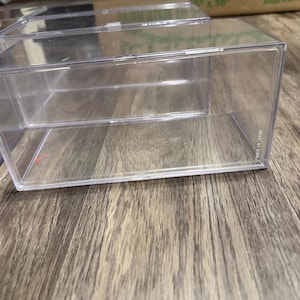 May include: A rectangular, clear acrylic display case with a removable lid. The container features transparent walls and a flat base, designed for organizing or showcasing small items. The structure is entirely see-through, highlighting its simple, functional design for storage or presentation purposes.