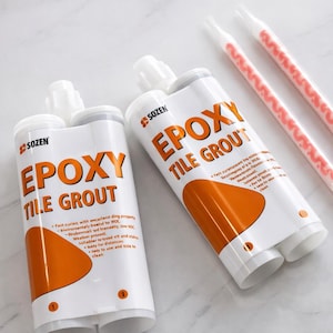 May include: Two white tubes of "Epoxy Tile Grout" with orange detailing and text. The tubes have white nozzles and are accompanied by two pink and white mixing tips. The product is labeled "SOZEN".