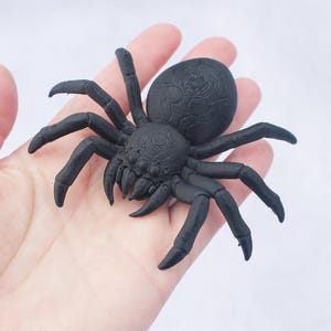May include: A black spider figurine with detailed legs and body, resting on a hand. The spider has a textured surface and is a dark, matte black color. The background is a plain white.