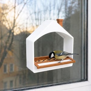 May include: A white house-shaped bird feeder with a brown chimney and a brown feeding tray attached to a window. A small bird with a black head, yellow chest, and grey wings is perched on the tray eating seeds from the feeder.