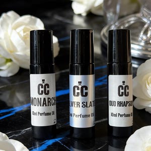 Puede incluir: Tres frascos de aceite de perfume con tapas negras y etiquetas. Las etiquetas dicen "MONARCH", "SILVER SLATE" y "OUD RHAPSODY". Cada botella tiene el logotipo "CC" y el texto "10ml Perfume Oil". Flores blancas en el fondo.