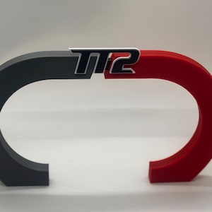Pode incluir: Uma escultura de dois tons com um design curvo preto e vermelho. A escultura apresenta as letras "TT2" em prata e preto. O design é uma representação moderna e abstrata.