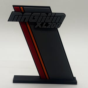 May include: A black and grey decorative sign featuring the text "Magnum XL200". The sign has a red and orange stripe on the side and sits on a black base. The sign is angled.