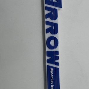 May include: A white and blue 3D-printed sign featuring the word "ARROW" in bold blue letters. The text "dynamics inc." is below in a smaller blue font. The sign is rectangular with a pointed base.
