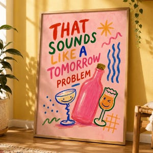 Könnte beinhalten: Ein gerahmter Kunstdruck auf rosa Hintergrund mit dem Text THAT SOUNDS LIKE A TOMORROW PROBLEM in mehrfarbiger Schrift. Die Illustration zeigt eine rosa Flasche und zwei Gläser mit lächelnden Gesichtern, umgeben von abstrakten Formen.