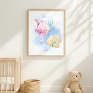 Op de afbeelding: Ingelijste aquarelprint met twee cartoonvissen. Eén is roze, de andere geel met blauwe strepen. De achtergrond heeft blauwe aquarelwashes en bellen, waardoor een onderwaterlandschap ontstaat.