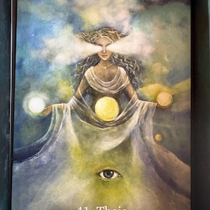 Può includere: Una carta oracolare con un'illustrazione ad acquerello di una donna in abito bianco, che tiene globi di luce. La carta ha uno sfondo blu e verde. Il testo sulla carta recita "41. Theia Divine Light Reveals All Truth."