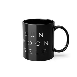 Op de afbeelding: Een zwarte keramische mok met de woorden "SUN MOON SELF" in het wit gedrukt. De mok heeft een gebogen handvat en een glanzende afwerking. Het ontwerp is eenvoudig en modern, geschikt voor koffie of thee.