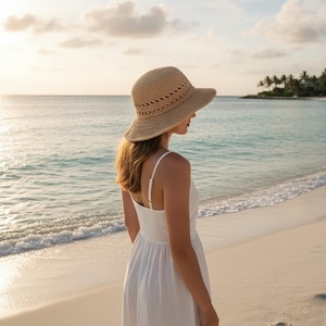 May include: A person wearing a beige woven sun hat with a wide brim and a white sundress stands on a sandy beach, looking out at the sea. The sky is a soft, warm colour, and the water is a light blue.