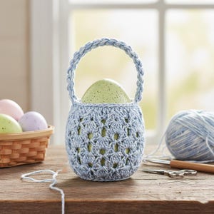 May include: A light blue crocheted Easter basket with a braided handle, holding a speckled green egg. A basket of pastel eggs sits to the left. A ball of blue yarn and crochet tools are on a wooden surface.