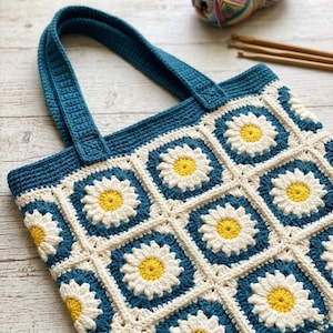 May include: A handmade crochet tote bag featuring a daisy pattern. The bag has a teal blue handle and trim, with white and teal blue squares forming the background. Each square has a yellow and white daisy in the center. The bag is made of yarn.