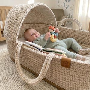 May include: A baby lies in a cream-coloured knitted bassinet, holding a fox and moon toy. The bassinet has a curved hood and braided handles. The baby wears a green outfit and striped socks. A small book is visible.