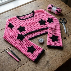May include: A bright pink crochet long-sleeve crop top featuring a black trim around the neckline, cuffs, and hem. The garment is decorated with several black crocheted star appliques scattered across the front and sleeves. The knit is open.