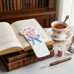 May include: A handmade white crochet bookmark shaped like a paw print, with pink and blue accents. The bookmark is placed in an open book, with a teacup and crochet hook nearby. The bookmark features a paw print design and a blue bow.