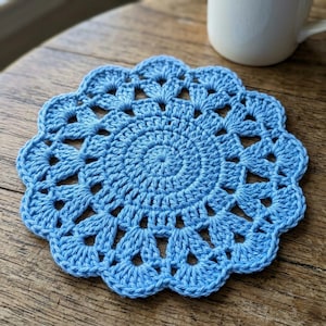 May include: A light blue, handmade crochet coaster with a scalloped edge. The coaster is circular with an open, lacy design. The coaster is sitting on a wooden surface, with a white mug in the background. The coaster is approximately 13 cm in diameter.