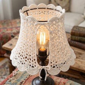 Può includere: Una lampada da tavolo in stile vintage con un paralume all'uncinetto color crema. Il paralume ha un bordo smerlato ed è legato con fiocchi. La lampada presenta una lampadina Edison decorativa e poggia su una base in metallo scuro.