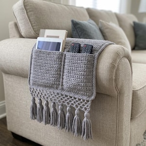 May include: A gray crocheted sofa arm organizer with two pockets and a fringe of tassels. The pockets hold a tablet, books, and two remote controls. The organizer is draped over the arm of a beige sofa.