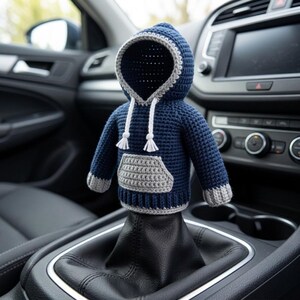 May include: A miniature crochet hoodie cover for a gear shift knob, featuring a dark blue body with a light gray front pocket, cuffs, and hem. The hood is trimmed in light gray and includes two white drawstrings with small tassels at the ends.