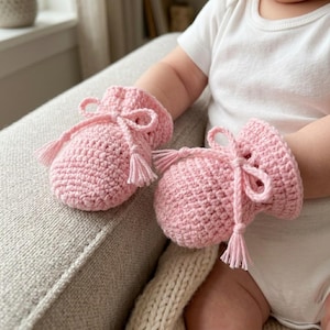 Op de afbeelding: Een paar lichtroze gehaakte babyslofjes. Elk slofje heeft een trekkoordsluiting met een strik en kwastjes. De slofjes zijn gemaakt van zacht, getextureerd garen en zijn ontworpen voor een pasgeborene.