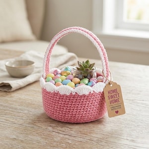 Puede incluir: Una cesta rosa de ganchillo con un borde y asa festoneados blancos, llena de caramelos de colores pastel y una pequeña suculenta en un tarro de cristal. Una etiqueta dice "Home Sweet Home, Handmade with Love."