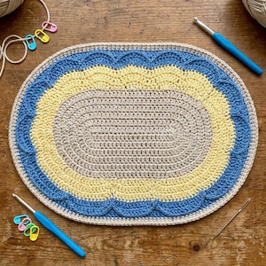 May include: An oval-shaped, handmade crochet rug with a beige center, surrounded by a yellow and blue scalloped border. The rug is placed on a wooden surface, with crochet hooks and stitch markers nearby. The rug measures approximately 24 inches long and 18 inches wide.