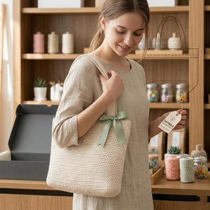 Pode incluir: Uma sacola tote bege de crochê com uma fita verde claro amarrada em um laço. A sacola é segurada por uma pessoa vestindo uma blusa bege. Uma etiqueta que diz "GIFTABLE FOR EVERYONE" está presa à sacola.