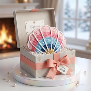 May include: A gift box with a crocheted seashell design in pink, blue, and white. The box is tied with a pink ribbon and a tag that says "From: Sarah". Inside the box is a gift card. The box is on a white surface.