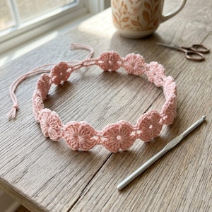 May include: A light pink crochet headband featuring a repeating pattern of small, six-petaled flowers connected in a circular chain. The headband includes two long, thin matching crochet ties at the ends for securing the piece in place.