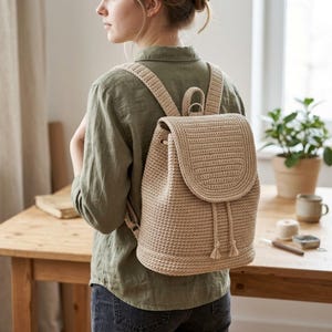 Puede incluir: Una mochila de ganchillo en color beige con cierre de solapa estructurado, cierre de cordón con borlas en los extremos y dos correas ajustables para los hombros. El bolso presenta un patrón de tejido denso y uniforme en tono arena.