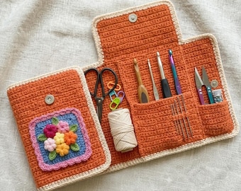 May include: An open orange crochet case with a cream border, containing crochet hooks, scissors, and other tools. The case features a square floral design on the front and a button closure. A ball of yarn is also present.