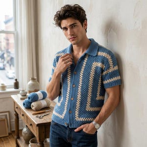 May include: A short-sleeved crochet shirt featuring a geometric pattern in blue and cream. The garment has a collared neckline, a button-down front with five visible brown buttons, and a textured knit construction. The shirt is paired with blue jeans.