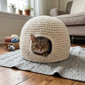 May include: A dome-shaped, off-white crocheted cat bed with a circular opening. A brown tabby cat is resting inside the structure. The bed is crafted from thick, chunky yarn with a visible stitch pattern, providing a cozy, enclosed space for a pet.
