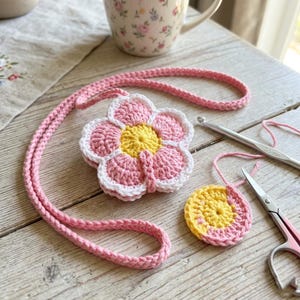 May include: A pink crochet flower necklace with a matching cord. The flower features pink petals, a yellow centre, and a white border. A crochet circle and crafting tools, including scissors and a 3.5 mm crochet hook, are also present.
