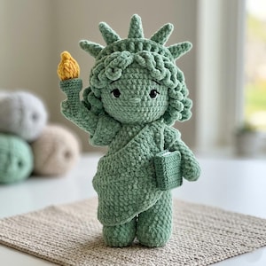 Op de afbeelding: Een gehaakte Statue of Liberty pop in groen, met een gele fakkel en een boek. De pop heeft een gedetailleerde kroon en krullend haar. Hij staat op een beige oppervlak. Een zachte sculptuur van het iconische monument, geschikt voor decoratie of als speelgoed.