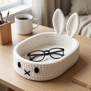 May include: A white, crocheted bunny-shaped basket with black embroidered eyes and mouth, holding a pair of black-framed glasses. The basket has two upright ears. A white mug of coffee and a wooden pen holder are in the background.